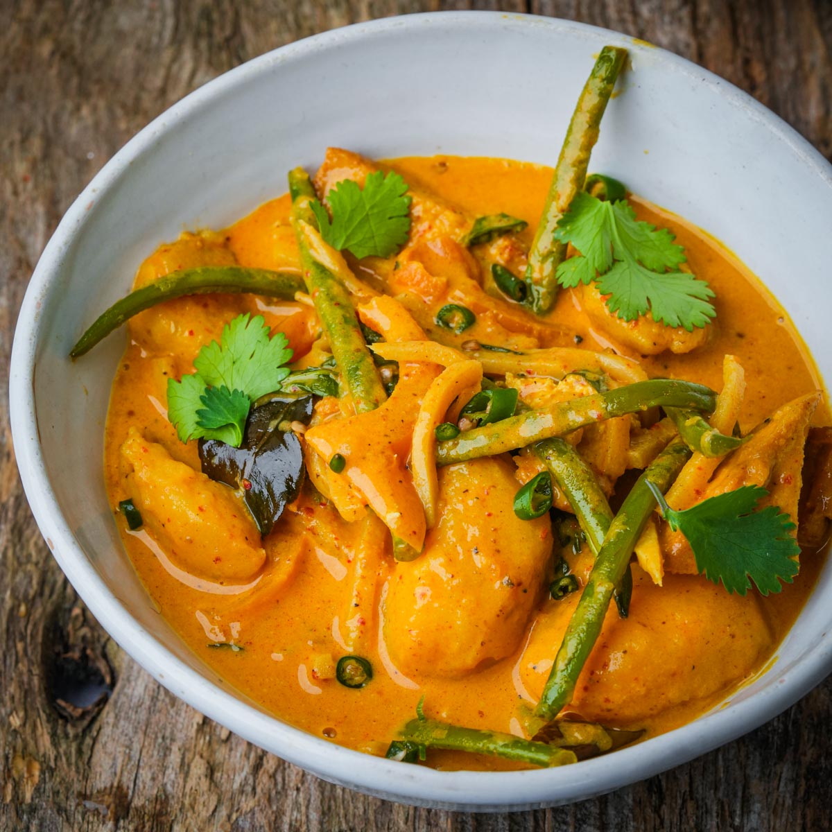 A bowl of vegan yellow curry with chunks of tofu, green beans, sliced onions, and cilantro leaves on a wooden surface.
