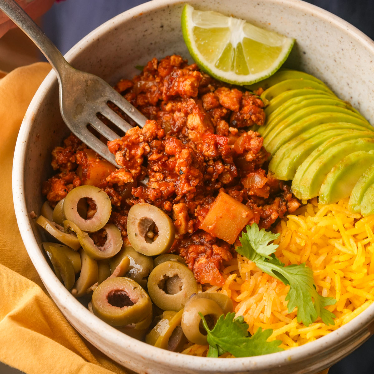 A bowl containing vegan picadillo, sliced avocado, yellow rice, sliced green olives, fresh cilantro, and a lime wedge, with a fork resting inside.